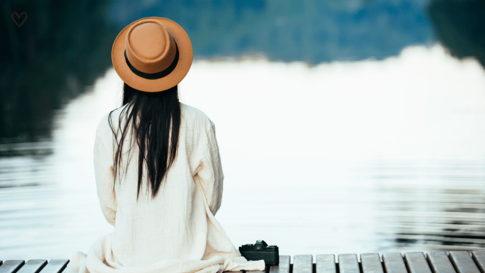 Žena sedící zády u klidné vodní hladiny, v tichém zastavení a spočinutí.